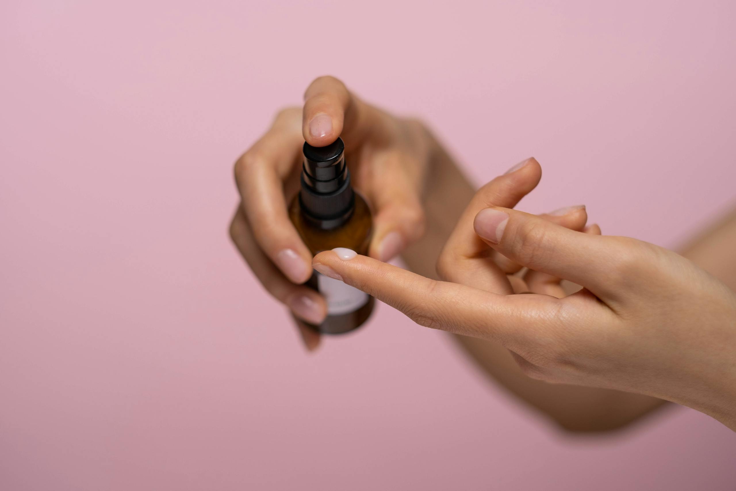 Hands applying skincare serum from a spray bottle against a pink background.